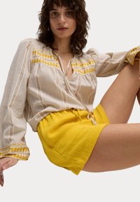 Blusa crema con detalles bordados en amarillo, de manga larga y rayas verticales, combinada con un short amarillo texturizado.