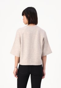 Pullover cu maneci scurte, de culoare bej, cu un guler rotund și un finisaj texturat. Prezintă mâneci raglan și un tiv scurt, asortat cu blugi negri.