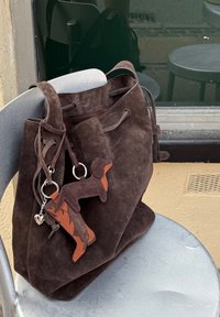 Brązowa torba typu bucket wykonana z miękkiego zamszu, z odpinanym breloczkiem przedstawiającym psa i but w ciemniejszym odcieniu brązu z pomarańczowymi akcentami.