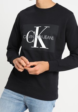 Osoba ubrana w czarną bluzę Calvin Klein Jeans z dużym białym logo „cK” na piersi i jasnoniebieskie dżinsy.