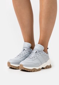 Svetle modré tenisky s hladkým koženým svrškem, kulatou špičkou a texturovanou gumovou podrážkou. Obsahují šněrování, polstrovaný límec a nenápadné logo.