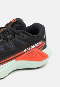 Chaussures de course noires avec tige en maille, accents orange et rouge, semelle texturée et talon vert menthe clair. Présente un logo bien visible et des détails réfléchissants.
