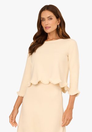 Beige, gerippter Oberteil mit Dreiviertelärmeln, der einen gewellten, fransigen Saum und Ausschnitt hat. Weicher Stoff und lässige Passform.