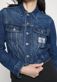 Blå denimjacka med knappstängning, bröstfickor och ett varumärkestagg. Har en klassisk krage och texturerad tyg.