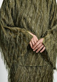 Fringed-textured oliivinvihreä vaate, jossa on kiiltäviä metallilankoja, leveät hihat ja avoimet cuffit, jotka esittelevät monimutkaista pinnan yksityiskohtaa.