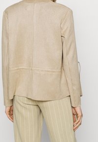Blazer beige perforé avec une coupe ajustée, présentant une texture lisse et des lignes épurées, associé à un pantalon clair à fines rayures.
