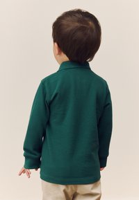 Žalia medvilninė ilgomis rankovėmis polo marškinėliai su tekstūruotu paviršiumi, gofruotu apykakliu ir šoninėmis skeltukėmis, derinami su šviesiai smėlio spalvos kelnėmis.