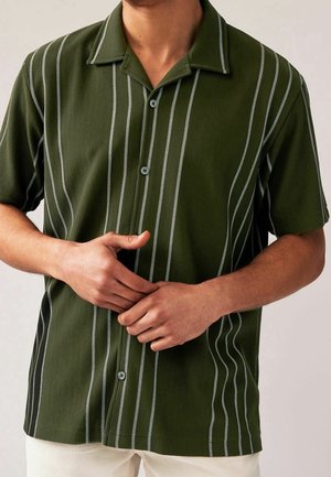 Mann trägt ein olivgrünes, kurzärmeliges Hemd mit weißen vertikalen Streifen, die Hände unter der Brust zusammengelegt, beige Hose sichtbar.