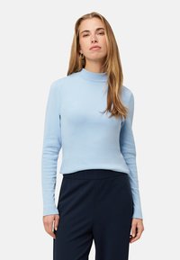 Haut à col roulé bleu clair à manches longues en tissu doux, présentant une coupe ajustée et des poignets côtelés, associé à un pantalon foncé.