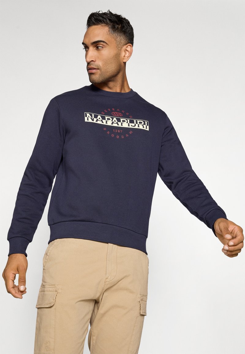 Homme portant un sweat à capuche bleu marine avec le logo Napapijri, un pantalon cargo beige, regardant vers sa gauche avec les bras légèrement étendus devant un fond uni.