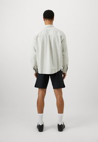 Gai pelēks garām piedurknēm krekls ar aizmugures jostu, salikts kopā ar melniem šortiem un melnām kurpēm, skatīts no aizmugures uz vienkārša fona.