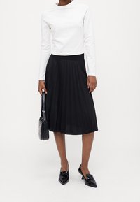 Vit långärmad topp, svart plisserad midi kjol, svarta lackskor och liten svart handväska med silverdetaljer.