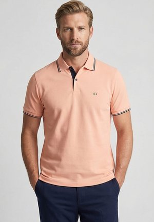 Man met baard die een perzikkleurig poloshirt met donkere rand en donkerblauwe broek draagt, staand met handen in de zakken tegen een effen achtergrond.