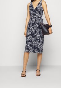 Kvinna i en marinblå ärmlös omlotklänning med vit blommönster, håller en svart handväska och har på sig svarta sandaler med remmar.