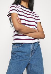 Camiseta de manga corta a rayas en blanco, azul marino y rojo, con cuello redondo y un ajuste relajado, combinada con unos pantalones vaqueros de denim azul de talle alto.