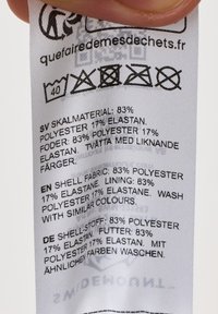 Vårdtagg i vitt tyg med svart text som anger materialkomposition: 83 % polyester, 17 % elastan; inkluderar skötselråd och ikoner.