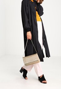 Czarny długi płaszcz, pomarańczowy sweter w prążki, różowe spodnie, czarne sandały na obcasie z otwartymi palcami oraz złota pikowana torebka na łańcuszku.