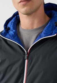 Uomo che indossa una giacca nera con cappuccio imbottito blu sopra una maglietta grigia a girocollo, la cerniera della giacca è parzialmente chiusa sul petto.