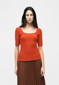 Γυναίκα με μακριά σκούρα μαλλιά, φορώντας εφαρμοστή πορτοκαλί μπλούζα με ριμπ ύφασμα και μανίκια μέχρι τον αγκώνα, καθώς και καφέ πλισέ φούστα, στέκεται μπροστά σε λευκό φόντο.