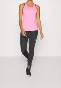 Różowy sportowy top na ramiączkach z wycięciem typu scoop, zestawiony z czarnymi legginsami. Legginsy mają subtelne logo po boku i gładką teksturę.