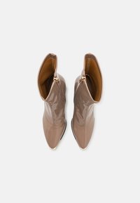 Paire de bottines taupe brillantes à bouts pointus, tirettes de fermeture éclair dorées et hauts repliés sur fond blanc.