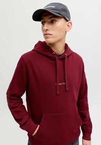 Maroon huppari, jossa on etutasku ja nyörivarrillinen huppu, korostettuna kultaisella brodeeratulla tekstillä. Yhdistetty tumman väriseen lippikseen.