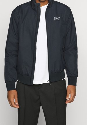 Mann trägt eine schwarze EA7 Emporio Armani Jacke mit Reißverschluss über einem weißen Hemd und schwarzen Hosen, die Hände entspannt an den Seiten.