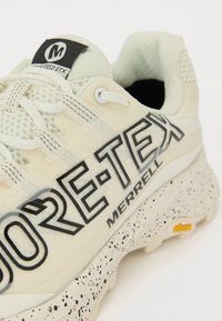 Lys beige Merrell turistsko sko med teksturert overdel, svart "GORE-TEX" merkevare og en prikkete hvit mellomsåle med Vibram yttersåle.