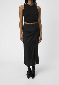 Un crop top noir à côtes associé à une jupe midi noire nouée avec des rayures texturées et une fente sur le côté, complété par des talons aiguilles en vernis.
