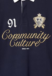 Marineblaues Poloshirt mit weißem Kragen, das einen großen goldenen "Community Culture"-Text, ein Wappenemblem und die Zahl "91" in Weiß aufweist.