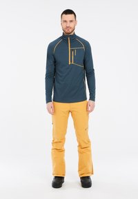 Mėlynas ilgomis rankovėmis viršus su oranžiniais akcentais ir užtrauktuku kišenėje, derinamas su ryškiai geltonomis kelnėmis. Lygus audinys, pritaikytas siluetas. Juodi batai.