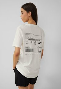 T-shirt en coton blanc avec un design postal noir imprimé au dos, comprenant du texte et des éléments graphiques, associé à un short noir.