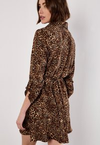 Robe en imprimé léopard en tissu doux, avec une taille cintrée, des manches longues et une coupe décontractée. Les couleurs incluent le marron et le beige avec des taches foncées.