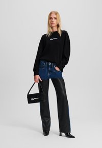 Črna majica s kapuco z belim logotipom, modri in črni jeans hlače s blokom barv ter majhna črna torbica. Model nosi špicaste črne čevlje.