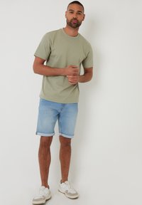 Mężczyzna stojący przy białej ścianie, ubrany w jasnozieloną koszulkę z krótkim rękawem, jasnoniebieskie podwinięte dżinsowe szorty i białe sneakersy.