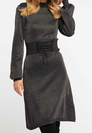Femme portant une robe noire pailletée à manches longues avec une taille froncée et un col montant, debout devant un fond uni.