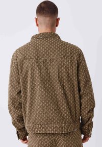 Homme portant une veste marron à motif et un pantalon assorti, vu de dos sur un fond gris clair.
