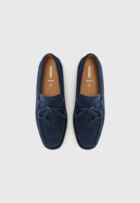 Mocassins en daim bleu marine avec un bout arrondi et un détail nœud sur le devant. Texture douce et doublure intérieure contrastante couleur cuir tanné. Marque Geox visible.