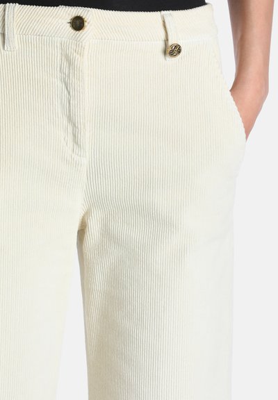 Gros plan sur un pantalon en velours côtelé crème à côtes avec des détails de boutons dorés et une main dans la poche droite sur fond blanc.