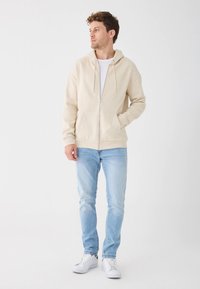 Felpa con zip color beige con coulisse, tasche a marsupio, abbinata a jeans azzurri chiari e sneakers bianche, in piedi su uno sfondo neutro.