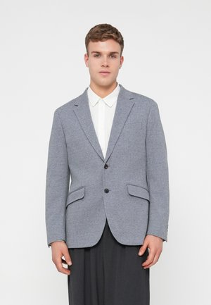Grauer strukturierter Blazer mit zwei Vorderaschen und einem einzelnen Knopfverschluss, getragen über einem weißen Hemd mit Kragen; taillierte Passform, modernes Design.