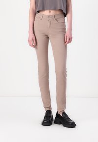 Jeans skinny beige avec une texture lisse, présentant un design classique à cinq poches et une fermeture à bouton à l'avant, portés avec des chaussures noires épaisses.