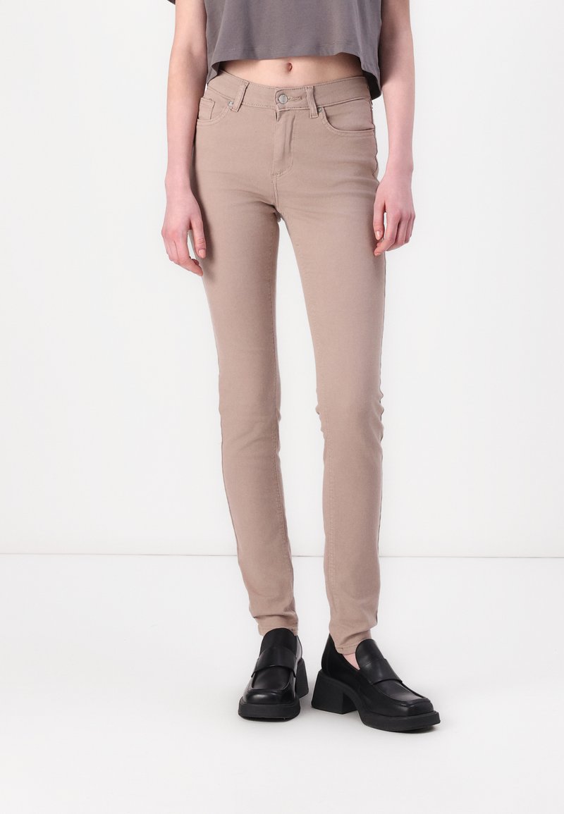 Jeans skinny beige avec une texture lisse, présentant un design classique à cinq poches et une fermeture à bouton à l'avant, portés avec des chaussures noires épaisses.