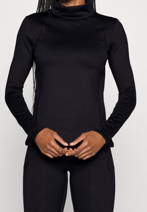 Personne aux longs cheveux tressés portant un col roulé noir moulant à manches longues et un pantalon noir assorti, les mains jointes à la taille.