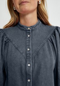 Graues Hemd mit Knopfleiste, hochgeschlossen, vertikaler Textur und gepufften Ärmeln. Verfügt über neun Knöpfe an der Vorderseite und ein Passe-Design.