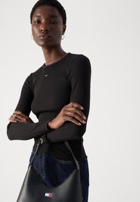 Black ribbed long-sleeve top with a round neckline, paired with dark denim. A black textured handbag features a small logo detail.