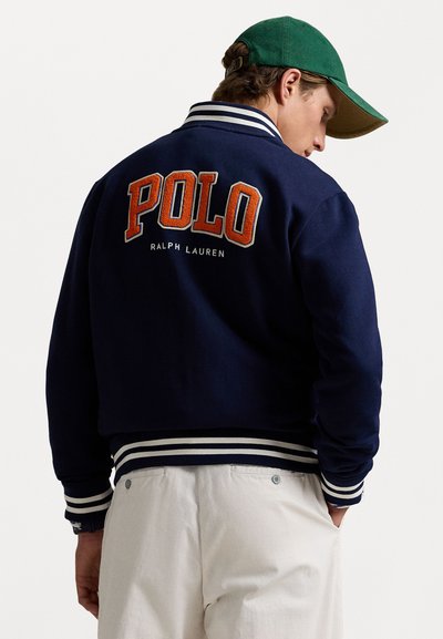 Námornícka bomber bunda s bielymi a modro pruhovanými manžetami, s veľkým oranžovým vyšívaním "POLO" na chrbte a "RALPH LAUREN" pod ním.