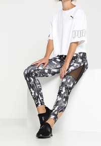 Bílý zkrácený tričko s logem Puma, v kombinaci s černobílými legínami s vzorem a síťovými výřezy a černými sportovními botami.