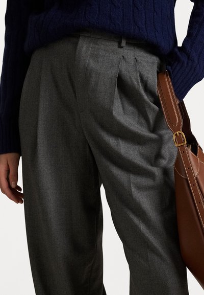 Graue, plissierte Hose aus einem leichten Stoff, kombiniert mit einem dunkelblauen Strickpullover. Eine braune Tasche mit goldenen Akzenten lehnt an der Hose.