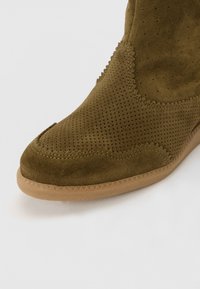 Bottine en suède marron avec texture perforée, détails cousus et semelle en caoutchouc tan avec un motif en zigzag sur le bord.